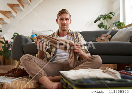 Portrait of confused man, sitting in living room with suitcase, holding pair of trousers, looking clueless, struggling to pack for holiday 113366596