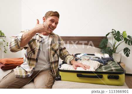 Portrait of handsome blond young man, going on vacation, recommending travel agency, encourages to go on holiday, sits on bed with suitcase, packs clothes luggage for travelling abroad Portrait of handsome blond young man, going on vacation, recommending travel agency, encourages to go on holiday, sits on bed with suitcase, packs clothes luggage for travelling abroad 113366901