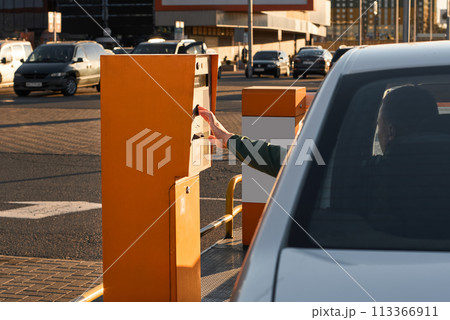 Man's hand take ticket in control tickets machine at vehicle entrance with boom barriers. Automatic barrier gates for supermarket parking lot. Security system for car park entry to shopping mall. 113366911