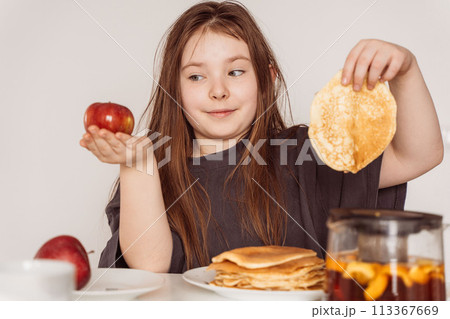 A little teenage girl holds a pancake in one hand and a red apple in the other and looks at it. Concept of choosing the right food. High quality photo A little teenage girl holds a pancake in one hand and a red apple in the other and looks at it. Concept of choosing the right food. High quality photo 113367669
