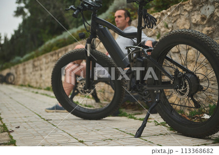 Selective focus on an innovated modern black electric bike, parked near a bench with a sitting cyclist 113368682