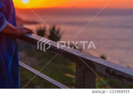 Handrail on which an elderly unrecognizable man leans with his hands stands against the background of an amazing sunset over the sea horizon, enjoying the magnificent landscape, view and life Handrail on which an elderly unrecognizable man leans with his hands stands against the background of an amazing sunset over the sea horizon, enjoying the magnificent landscape, view and life 113369353