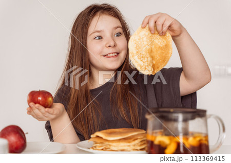 A little teenage girl holds a pancake in one hand and a red apple in the other and looks at it. Concept of choosing the right food. High quality photo A little teenage girl holds a pancake in one hand and a red apple in the other and looks at it. Concept of choosing the right food. High quality photo 113369415