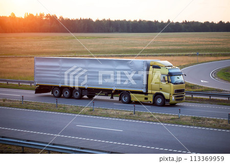 A truck with a tilt semi-trailer carries cargo along the highway in the evening against the backdrop of a summer sunset. The concept of cargo transportation, the logistics company, the work and rest 113369959