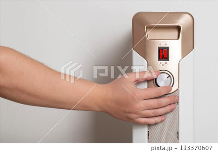 A man's hand regulates the heating power of an electric air heater in an apartment, close-up. Copy space for text, radiator 113370607