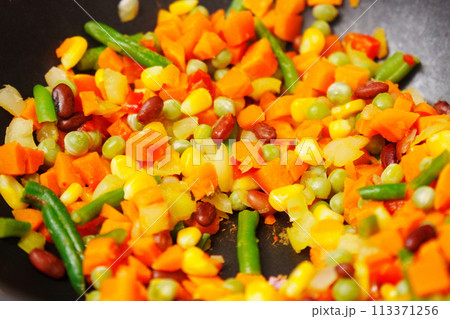 Colorful chopped vegetables is being stirred using a wooden spoon. The vegetables sizzle and release aromatic scents as they cook. 113371256