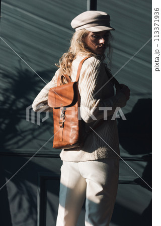 Studio portrait of beautiful woman with a curly blond hair holding brown backpack, posing on gray background. 113371936