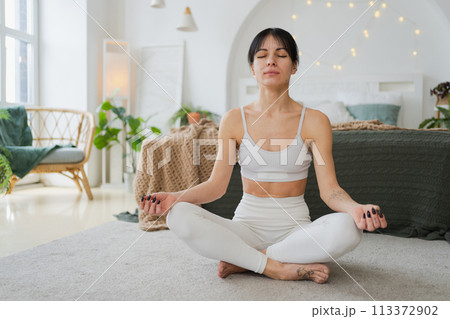 Yoga mindfulness meditation. Young healthy woman practicing yoga in living room at home. Woman sitting in lotus pose meditating smiling relaxing indoor. Girl doing breathing practice. Yoga at home 113372902