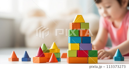 House built of colorful wooden children's cubes. Little baby is sitting on the floor of blurred background with copy space in white bright living room 113373273