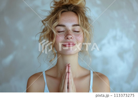 Blonde girl with curly hair doing in meditation the day with eyes closed, hands in prayer position, her face is calm and peaceful 113373275