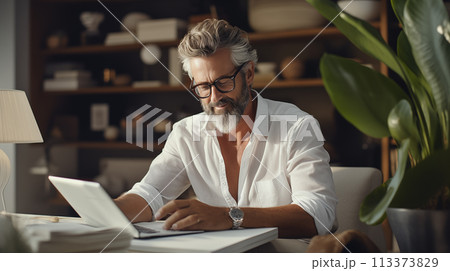 A stylish man with a beard is sitting at a table using a laptop in a building A stylish man with a beard is sitting at a table using a laptop in a building 113373829