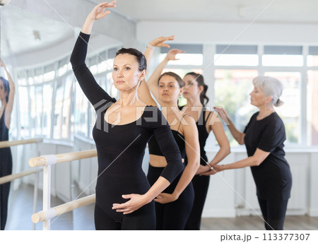Mature teacher conducts class with girls in ballet studio, observes doing forth position exercise. 113377307