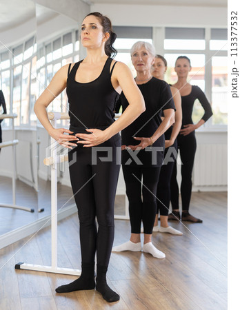 Women stand in fifth position near ballet barre during group training in dance studio Women stand in fifth position near ballet barre during group training in dance studio 113377532