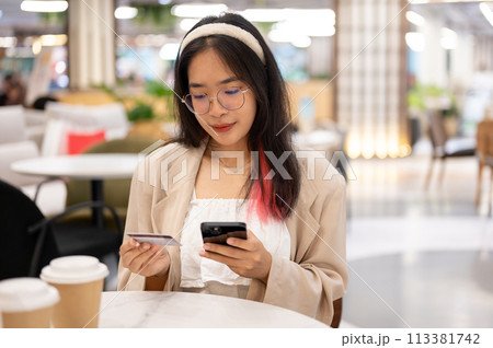 A woman is holding her smartphone and a credit card while sitting in a cafe in the shopping mall. 113381742