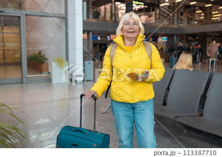 Airport terminal. Happy traveling Caucasian 60s woman is waiting at the departure gate to board the plane, using mobile smartphone and checking destination online.  113387710