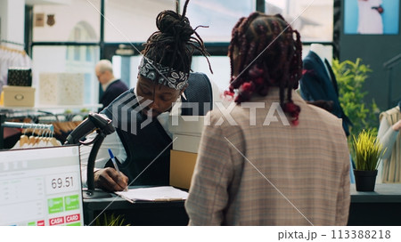 African american deliveryman receiving requested clothes packed in boxes, arriving at fashion showroom to pick up order and sign papers. Woman employee preparing items at cash register. Camera A. 113388218