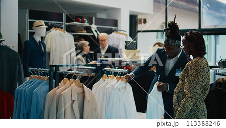 African american worker showing white shirts to pregnant woman, helping her to choose the right size to buy on discount. Store assistant recommending clothes from designer brands. Camera B. 113388246