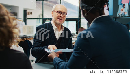 Retail assistants selling clothes to elderly client at fashion boutique, giving discount coupon to regular customer. Employees team assisting senior man to buy clothing items on sale. Camera B. 113388376