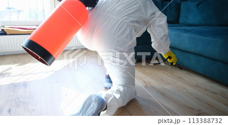 Worker in protective suit treating with disinfectant solution on floor under sofa in apartment Worker in protective suit treating with disinfectant solution on floor under sofa in apartment 113388732