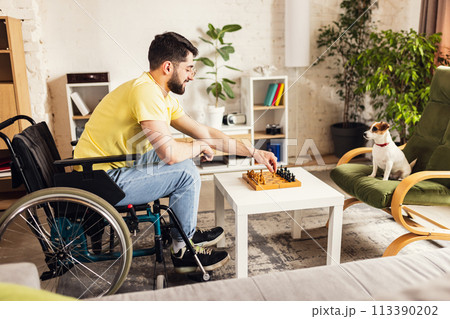 Smiling young man sitting on wheelchair in living room at home and playing chess with his little dog. Leisure time Smiling young man sitting on wheelchair in living room at home and playing chess with his little dog. Leisure time 113390202