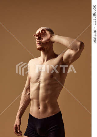 Shirtless muscular man shielding eyes from light against beige studio background. Young model with toned body. Shirtless muscular man shielding eyes from light against beige studio background. Young model with toned body. 113396650