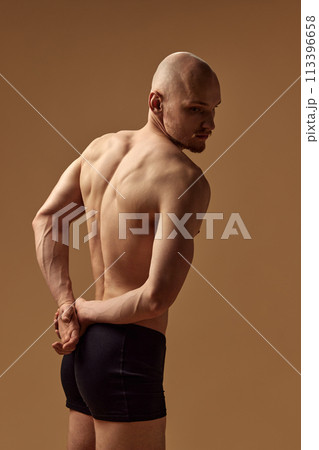 Shirtless young bald man holding hands behind back and showing his muscular back against beige studio background 113396658