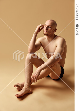 Shirtless man with perfect body curves posing with thoughtful expression in underwear on floor against beige studio background 113396677