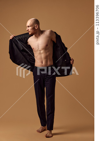 Shirtless young toned man dressed elegant trousers and jacket showing casual sophistication against sandy studio background. Shirtless young toned man dressed elegant trousers and jacket showing casual sophistication against sandy studio background. 113396706