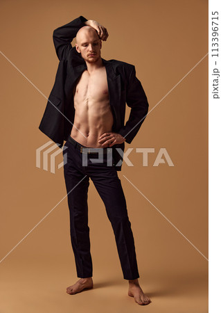 Shirtless young athletic man with elegant trousers and jacket showing casual sophistication against sandy studio background. Shirtless young athletic man with elegant trousers and jacket showing casual sophistication against sandy studio background. 113396715