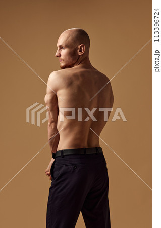 Rear view of shirtless man holding hands behind back and showing his muscular back against sandy studio background. Rear view of shirtless man holding hands behind back and showing his muscular back against sandy studio background. 113396724