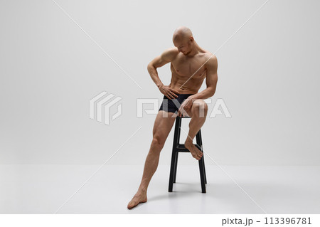 Athletic male model posing naked wearing in black underwear sitting on stool against grey studio background. Athletic male model posing naked wearing in black underwear sitting on stool against grey studio background. 113396781