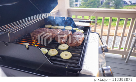 The outdoor two-burner gas grill is put to good use, sizzling with the sound and aroma of ribeye steaks and onion rings being perfectly cooked. The outdoor two-burner gas grill is put to good use, sizzling with the sound and aroma of ribeye steaks and onion rings being perfectly cooked. 113398875