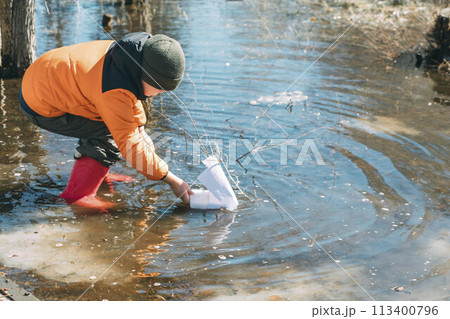 Springtime Sailor : Amid the budding trees, a young boy leans into the experience of launching a paper boat, symbolizing new beginnings and simple joys 113400796
