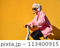 Smiling happy cool gray haired old senior woman on scooter or bicycle wearing headphones with a microphone. The Hands free concept 113400915