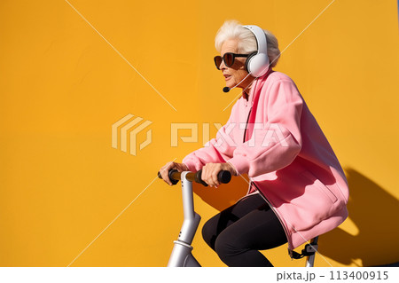 Smiling happy cool gray haired old senior woman on scooter or bicycle wearing headphones with a microphone. The Hands free concept Smiling happy cool gray haired old senior woman on scooter or bicycle wearing headphones with a microphone. The Hands free concept 113400915