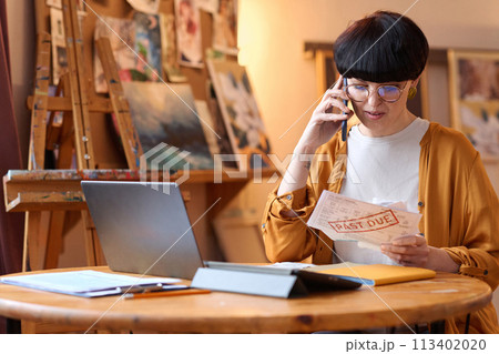 Portrait of woman holding Past due bill and speaking on phone in art studio, copy space 113402020