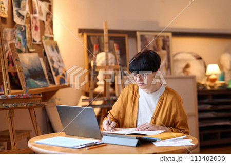 Wide angle view of funky female artist using laptop in art studio and taking notes while managing small business Wide angle view of funky female artist using laptop in art studio and taking notes while managing small business 113402030