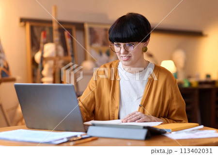 Portrait of smiling female artist using laptop in studio giving online art class and speaking via video chat 113402031