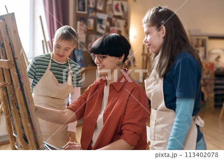 Side view portrait of smiling young woman painting on easel in art class with children watching Side view portrait of smiling young woman painting on easel in art class with children watching 113402078
