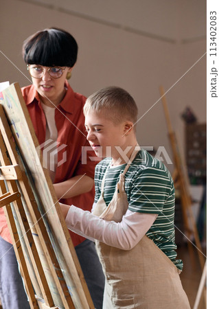 Vertical portrait of smiling boy with disability painting on canvas in art class and wearing apron 113402083