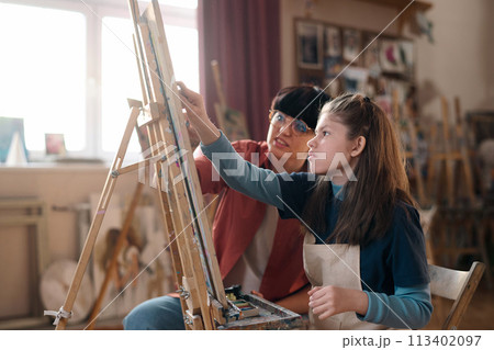 Side view portrait of young girl with disability painting on easel in art class with female teacher assisting copy space Side view portrait of young girl with disability painting on easel in art class with female teacher assisting copy space 113402097