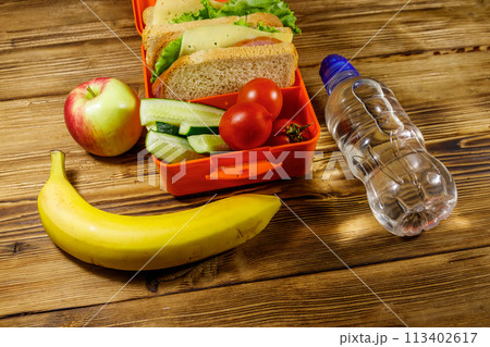 Bottle of water, apple, banana and lunch box with sandwiches and fresh vegetables on a wooden table Bottle of water, apple, banana and lunch box with sandwiches and fresh vegetables on a wooden table 113402617