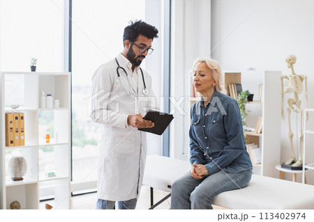 Bearded male doctor and elderly female patient sitting at exam couch. Bearded male doctor and elderly female patient sitting at exam couch. 113402974