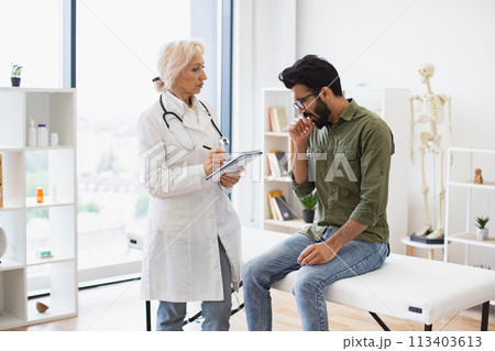 Caucasian senior female doctor and young male patient sitting at exam couch. Caucasian senior female doctor and young male patient sitting at exam couch. 113403613