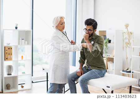 Elderly lady using stethoscope while young man sitting on exam couch. Elderly lady using stethoscope while young man sitting on exam couch. 113403623