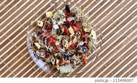 Salad of fresh vegetables and microgreens. Tomatoes, onions, apples, a mound of sprouted mung beans, sesame seeds, flax and cheese cubes on on a transparent plate on the table. Close-up 113406007