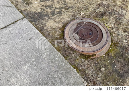 A rusty iron hatch with a pattern is located next to a dry grass pedestrian road during the daytime. Close-up 113406017