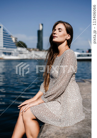 A woman with long hair is sitting on a dock by the water. She is wearing a white dress and she is looking at the water. The scene has a calm and peaceful mood. 113406066