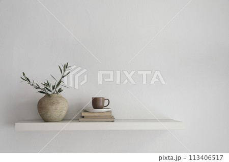 Elegant home still life. Floating shelf. Textured vase with green olive tree branches and old books. Cup of coffee, tea. Modern Mediterranean appartement. White wall background. Interior indoor mockup 113406517