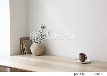 Breakfast, scandi indoor interior still life. Minimal home design concept. Beige ceramic vase with olive tree branches. Cup of coffee, tea on wooden table, desk with old books, empty beige wall mockup 113406518